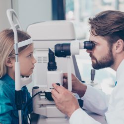 Health care, medicine, eye sight and technology concept. Focused brunet bearded optometrist with non contact tonometer is checking small blond`s girl patient intraocular pressure at eye clinic